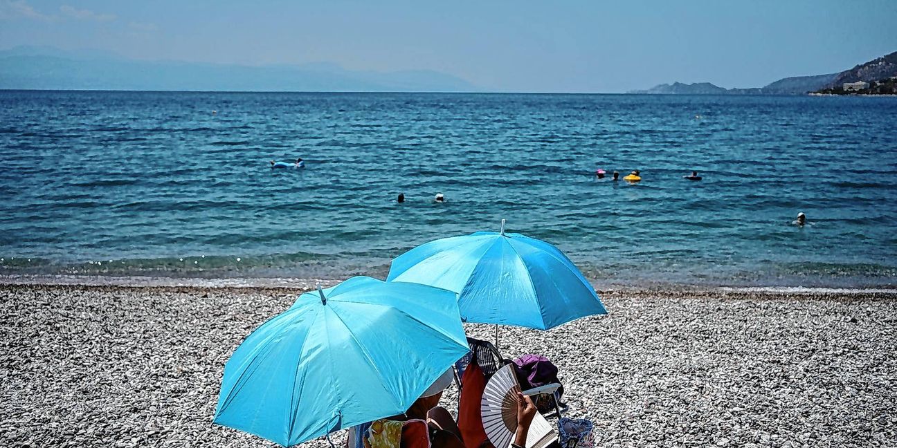 Eine Frau mit einem Handventilator unter einem Sonnenschirm am Strand.